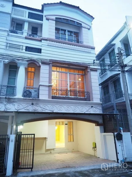 Townhouse for rent in Lat Phrao, Bangkok - Three-storey townhouse exterior at dusk with large windows, balconies and a gated carport in Lat Phrao, Bangkok.