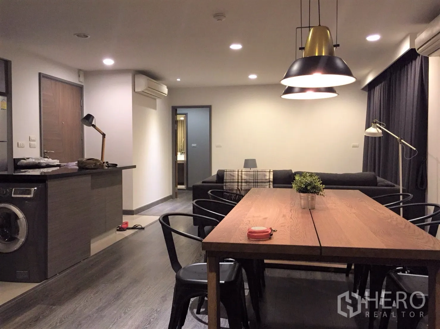 公寓 出租或出售在Watthana, Bangkok - 开放式客餐厅，木地板，长餐桌与吊灯，带洗衣机的厨房台面和沙发通向卧室。