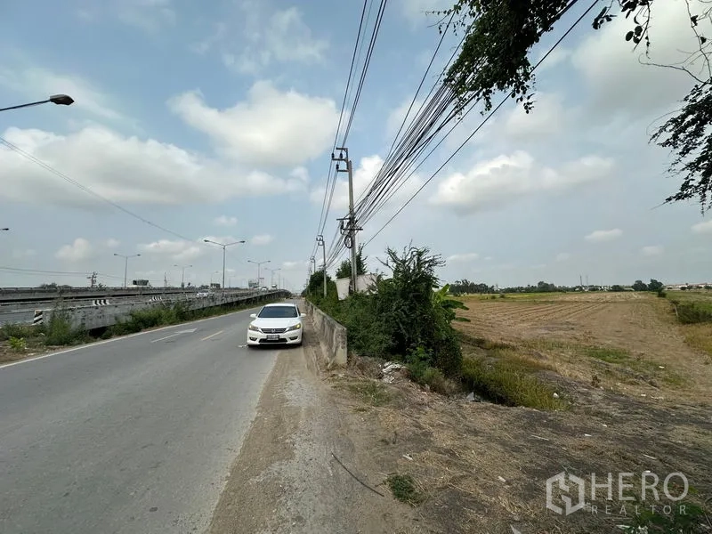 ที่ดิน ขายในBang Bua Thong, Nonthaburi - ถนนสองเลนมีรถจอด สายไฟพาดผ่าน และแปลงที่ดินกว้างทอดยาวทางขวา