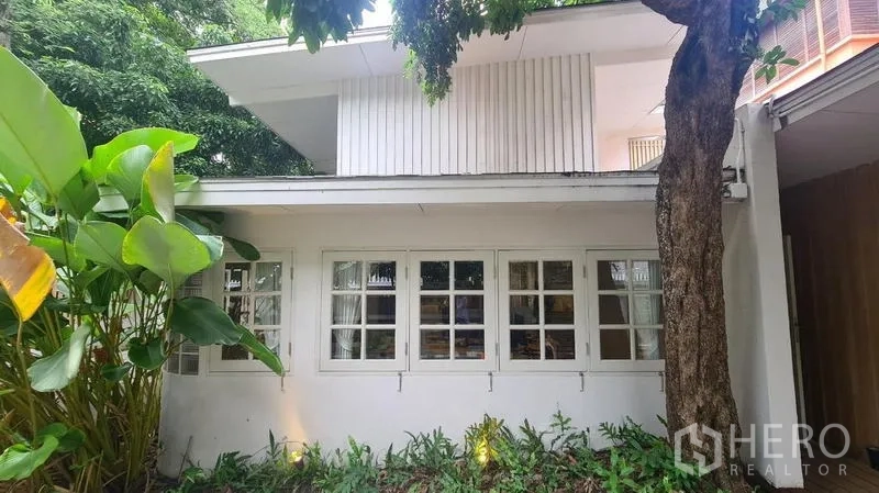 Detached House for rent in Watthana, Bangkok - White facade with grid windows framed by lush greenery.