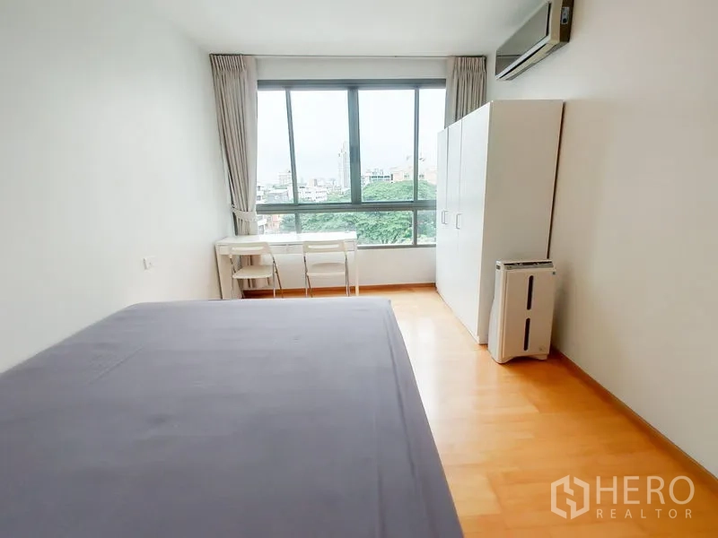Condominium for rent in Khlong Toei, Bangkok - Bedroom with large window, desk set and wardrobe overlooking greenery and city buildings.