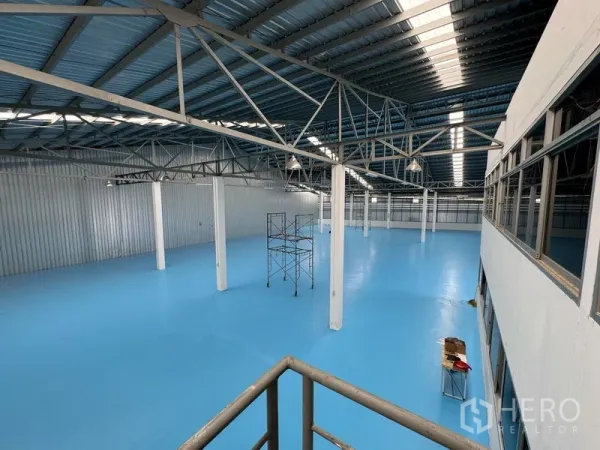 Warehouse for rent in Mueang Pathum Thani, Pathum Thani - Open warehouse interior with blue epoxy flooring, steel truss roof, skylights, and support columns.