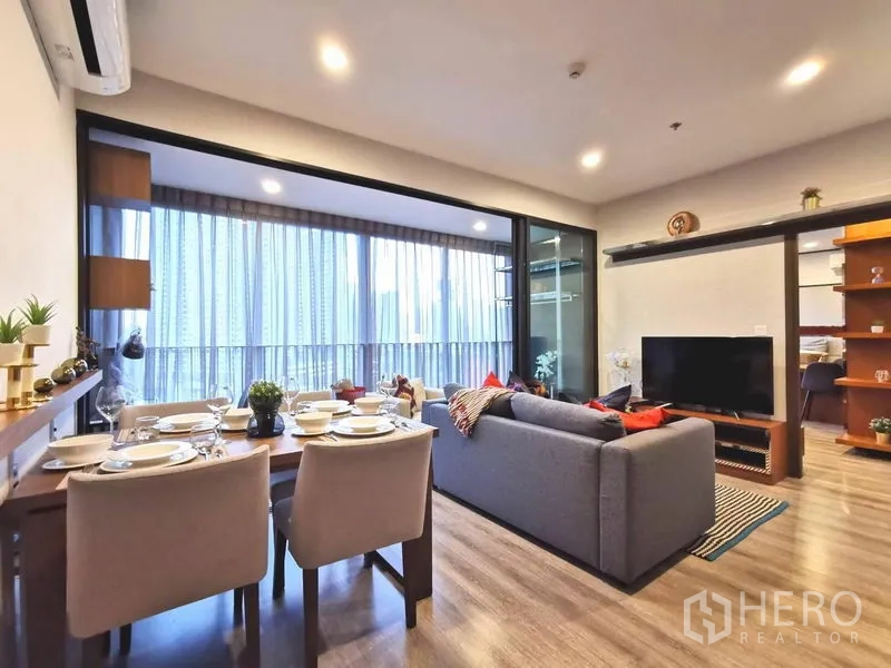 คอนโด ให้เช่าในHuai Khwang, Bangkok - พื้นที่นั่งเล่นและทานอาหารแบบโอเพนพร้อมโซฟาสีเทา โต๊ะทานอาหาร หน้าต่างเต็มบาน และระเบียงวิวเมืองในคอนโดกรุงเทพฯ