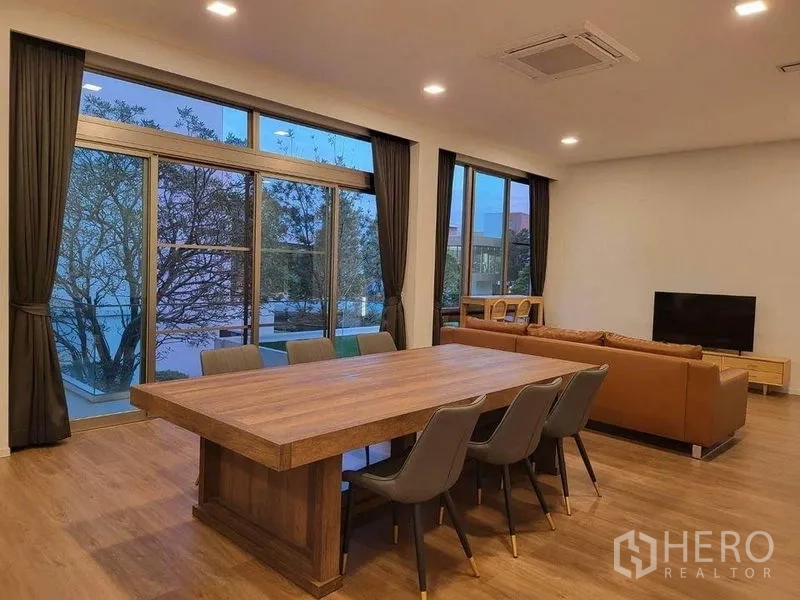 Detached House for rent in Saphan Sung, Bangkok - Open‑plan dining and lounge area with long wooden dining table, leather sofa and wide glass doors to the outdoors.