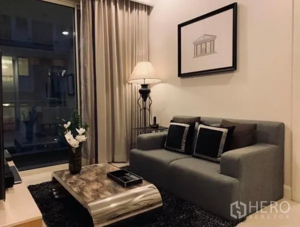 Condominium for rent in Pathum Wan, Bangkok - Cozy living corner with gray sofa, metallic coffee table, floor lamp, framed artwork, rug, and balcony access through sliding glass.