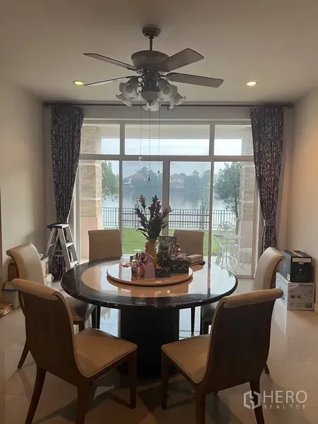 Detached House for rent or sale in Lat Krabang, Bangkok - Dining area with round table and ceiling fan opening to a lakefront garden through large glass doors.