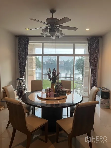 Detached House for rent or sale in Lat Krabang, Bangkok - Dining area with round table and ceiling fan opening to a lakefront garden through large glass doors.