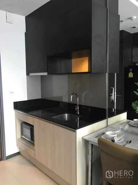 Condominium for rent in Watthana, Bangkok - Compact kitchenette with black stone countertop, sink, microwave, and dark cabinets.