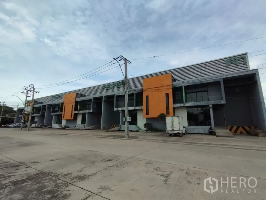 Factory for rent in Samut Prakan - Modern row of warehouse and factory units with orange facade panels, loading bays, and wide access road.