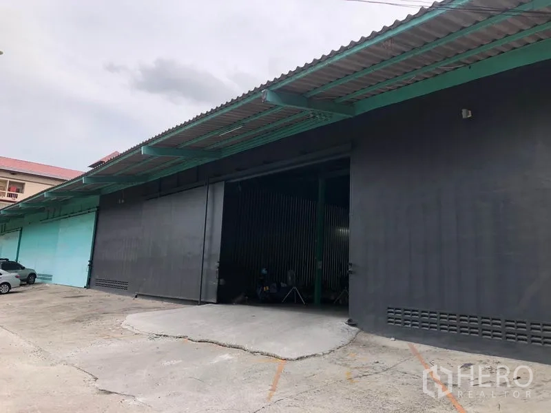 Warehouse for rent in Khan Na Yao, Bangkok - Front exterior of the warehouse with wide sliding metal doors, covered loading bay and a sloped driveway.
