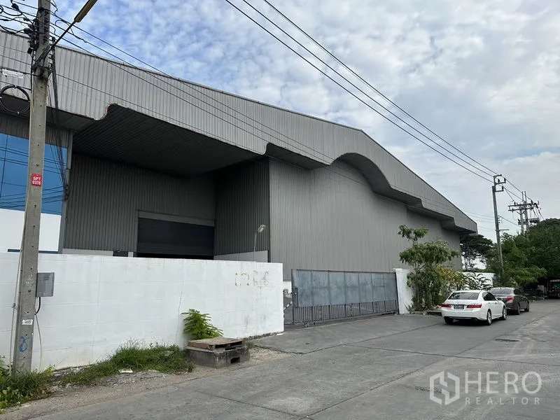 Warehouse for rent in Mueang Samut Prakan, Samut Prakan - Street-side view of corrugated steel warehouse with gated entrance, white perimeter wall and roadside parking.