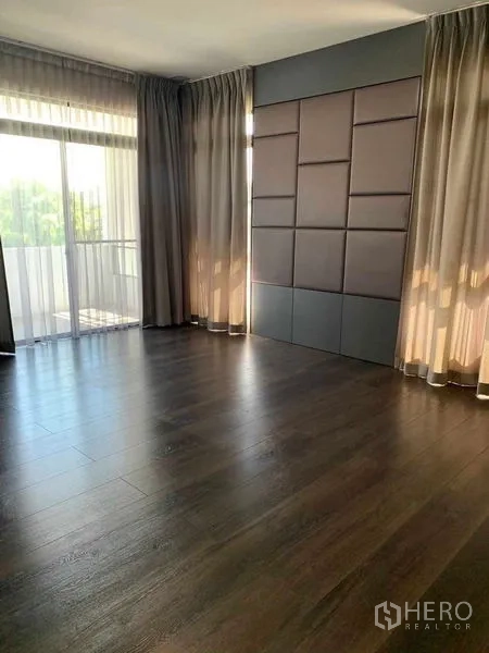 Detached House for rent or sale in Bang Phli, Samut Prakan - Spacious empty room with dark wood floor, feature panel wall and sliding glass door to a balcony.