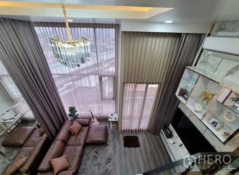 公寓 出租在Chatuchak, Bangkok - 挑高客厅，落地大窗与纱帘，水晶吊灯、棕色皮沙发、壁柜与城市景观。