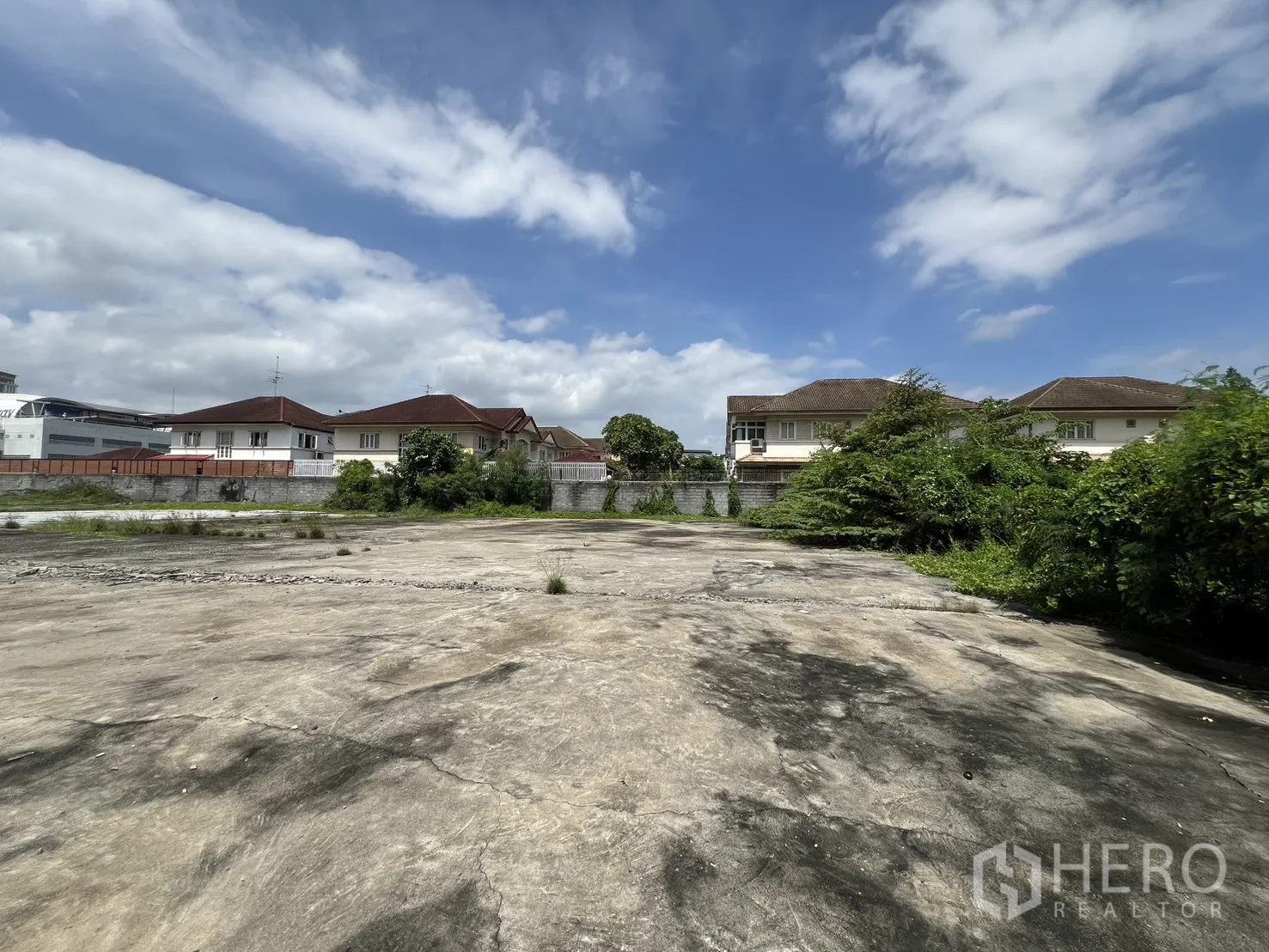 Land for sale in Bang Kapi, Bangkok - Open cleared plot with boundary wall and suburban houses at the rear under a blue sky.