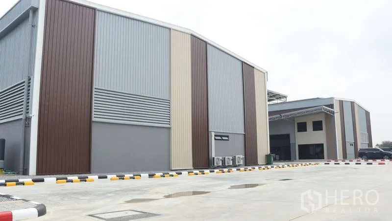 Warehouse for rent in Sam Phran, Nakhon Pathom - Angle view of metal‑clad warehouse façade with ventilation panels and parking area.