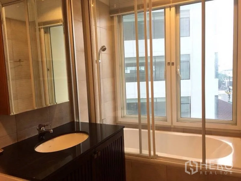 Condominium for rent in Watthana, Bangkok - Bathroom with bathtub beside big windows, black vanity counter and wall mirror.
