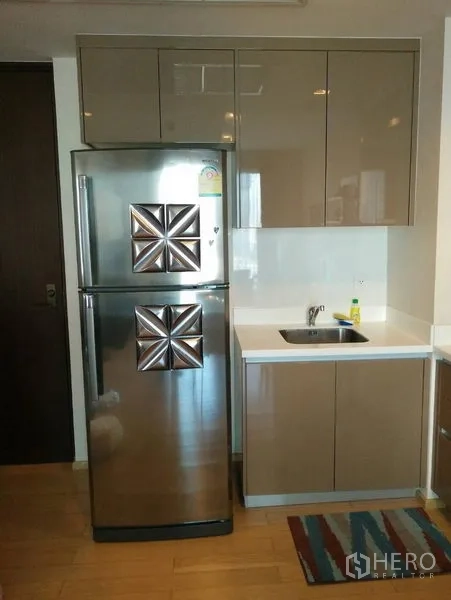 Condominium for rent in Khlong Toei, Bangkok - Compact kitchenette with glossy cabinets, stainless double-door fridge, sink and wood flooring.