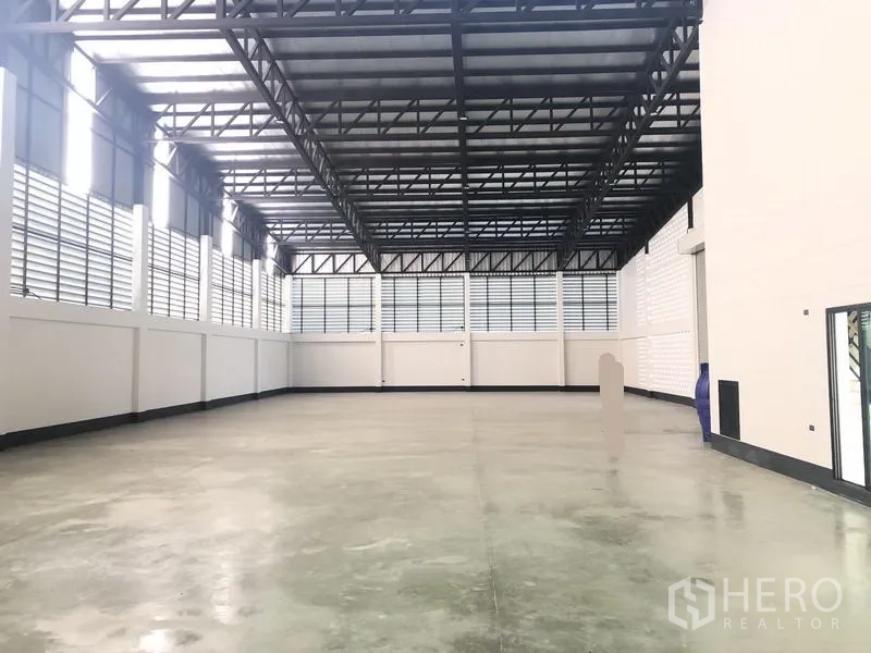 工厂 出售在Pathum Thani - 宽敞仓库内部，高钢桁架屋顶、抛光混凝土地面与百叶窗通风。