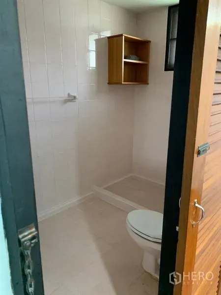 Detached House for rent in Khlong Toei, Bangkok - White-tiled bathroom with toilet, open shower zone and a small wooden shelf.
