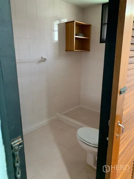 Detached House for rent in Khlong Toei, Bangkok - White-tiled bathroom with toilet, open shower zone and a small wooden shelf.