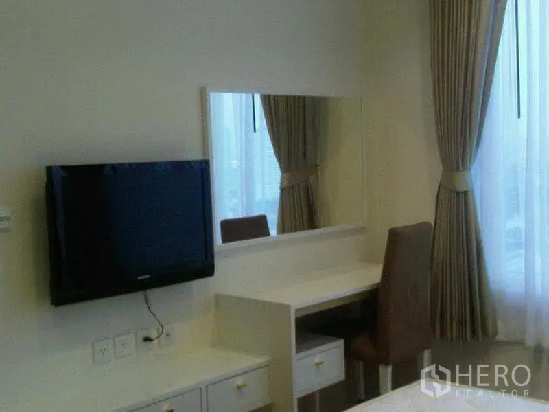 Condominium for rent in Khlong Toei, Bangkok - Bedroom corner with wall-mounted TV, dressing table with large mirror and tall curtains.