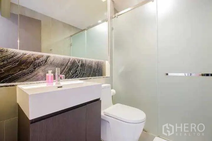 Condominium for rent in Khlong Toei, Bangkok - Modern bathroom with floating vanity, large mirror and frosted-glass shower enclosure.
