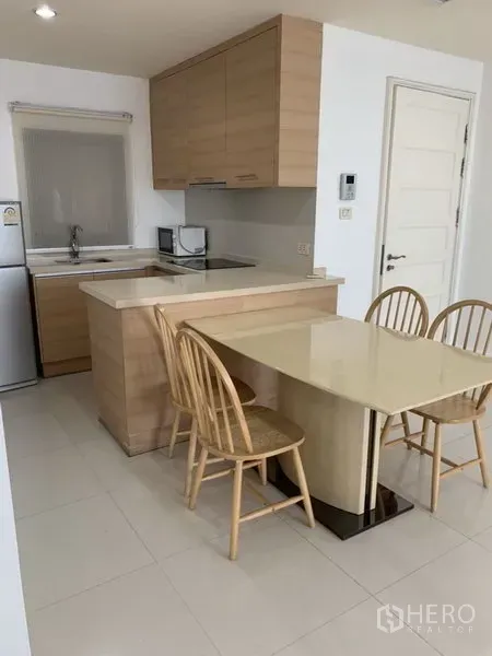 Condominium for rent or sale in Khlong Toei, Bangkok - Compact kitchen with light wood cabinets, breakfast bar, fridge and a four-seat dining table.