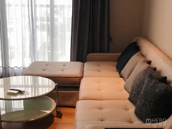 Condominium for rent in Khlong Toei, Bangkok - Neutral-tone sofa with ottoman and round glass coffee table beside tall window.