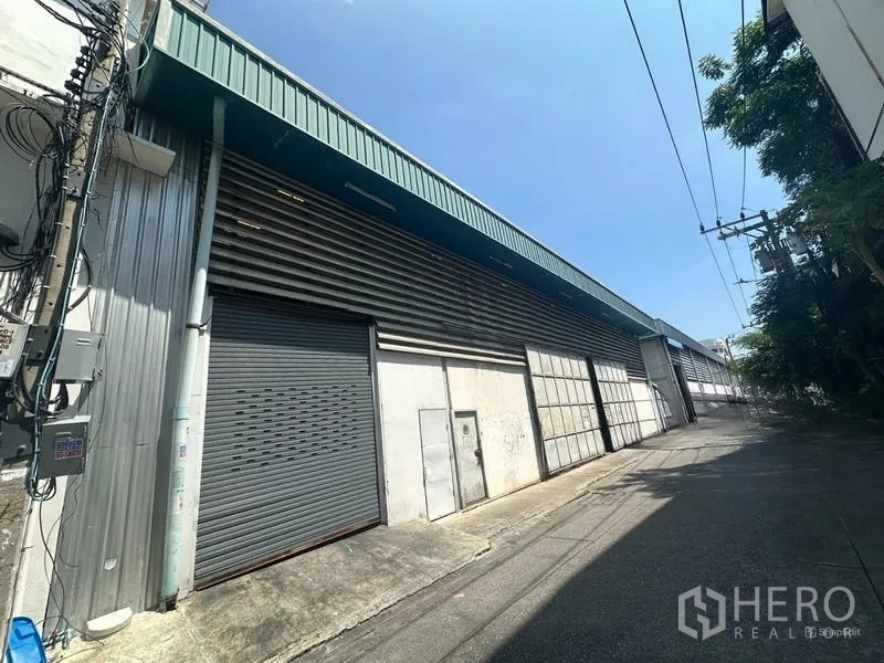 Warehouse for rent in Suan Luang, Bangkok - Street-side view of a long metal-clad warehouse with roller shutter and sliding gates in Suan Luang, Bangkok.