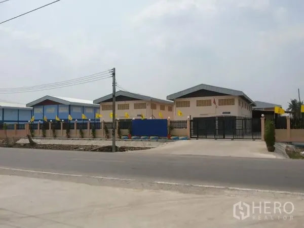 Factory for rent in Lat Lum Kaeo, Pathum Thani - Gated factory compound with multiple warehouse buildings and a wide concrete entrance along a paved road in Lat Lum Kaeo.