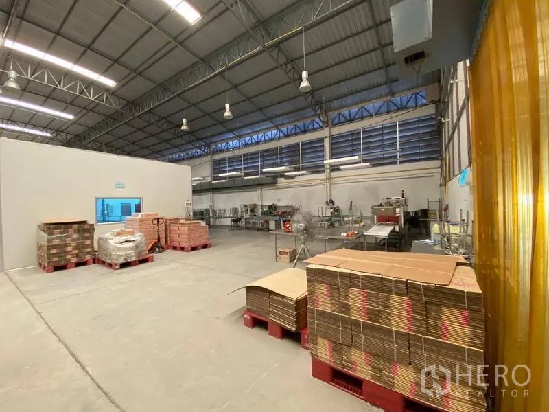 Warehouse for rent in Bueng Kum, Bangkok - Spacious storage zone with cardboard on pallets, machinery, and high louvered windows.