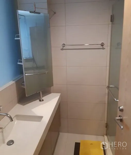 Condominium for rent in Khlong Toei, Bangkok - Tiled bathroom with long vanity, wall mirror, glass-enclosed shower and chrome fittings.