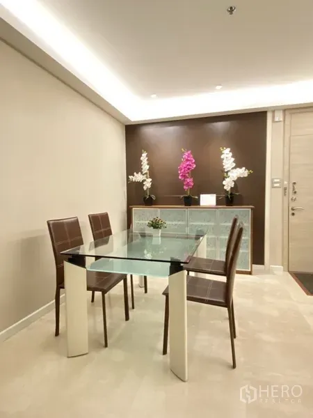 Condominium for rent in Bang Rak, Bangkok - Dining area with glass table and four chairs beside a feature wall and soft ceiling lights.
