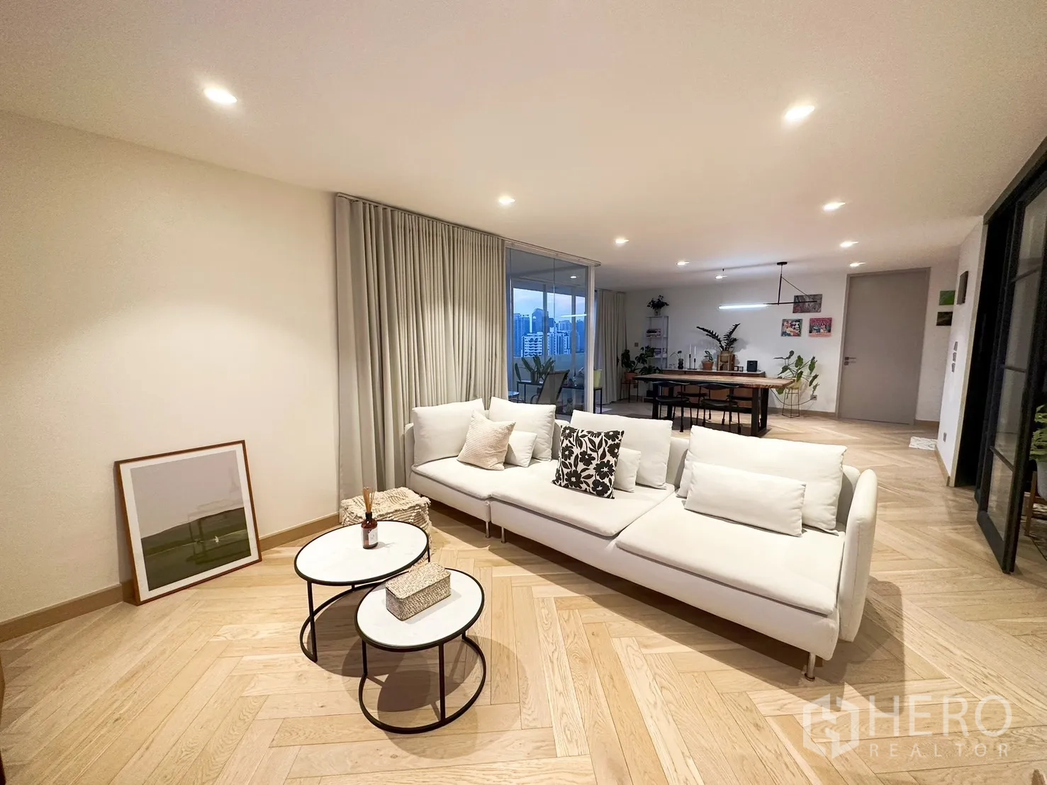 คอนโด for rent or sale in Watthana, Bangkok - ห้องนั่งเล่นโอเพ่นพร้อมโซฟาสีขาว พื้นไม้ลายก้างปลา และผนังกระจกเชื่อมโซนทานอาหารวิวเมือง.