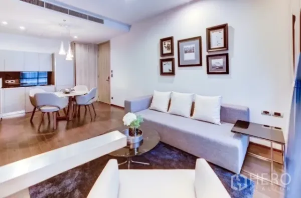 Condominium for rent or sale in Watthana, Bangkok - Open-plan living and dining area with a gray sofa, framed wall art, pendant lights, wood floors, and a compact fitted kitchen.