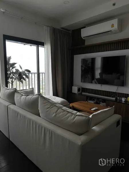 Condominium for rent in Khlong Toei, Bangkok - Bright living room with white sectional sofa, built-in TV wall and balcony door in Bangkok condo.