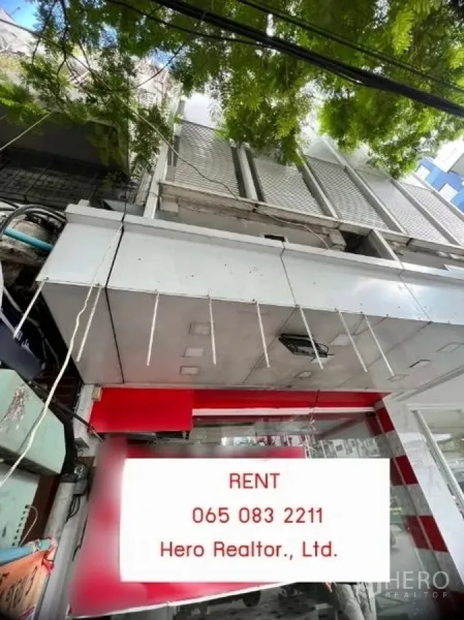Shophouse for rent in Khlong Toei, Bangkok - Street-facing shophouse exterior with glass frontage, metal awning, and roll-up shutter beneath shade trees in the Nana area.