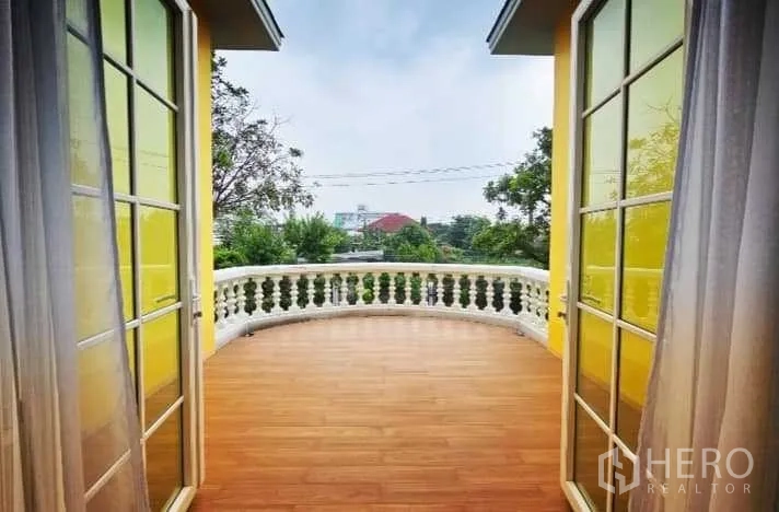 独立式住宅 出售在Phra Khanong, Bangkok - 开放露台，白色栏杆，两侧黄色墙面，远眺树木与社区景观。