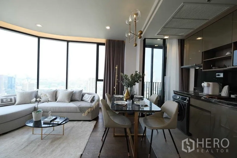 Condominium for rent in Khlong Toei, Bangkok - Corner living-dining space with panoramic windows, curved sofa, and sleek kitchen.