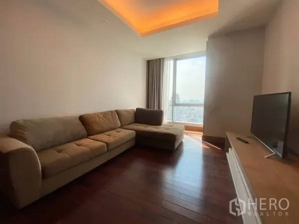 Condominium for rent in Sathon, Bangkok - Bright living room with L-shaped sofa, wood floors, and a floor-to-ceiling window showing Bangkok skyline views.