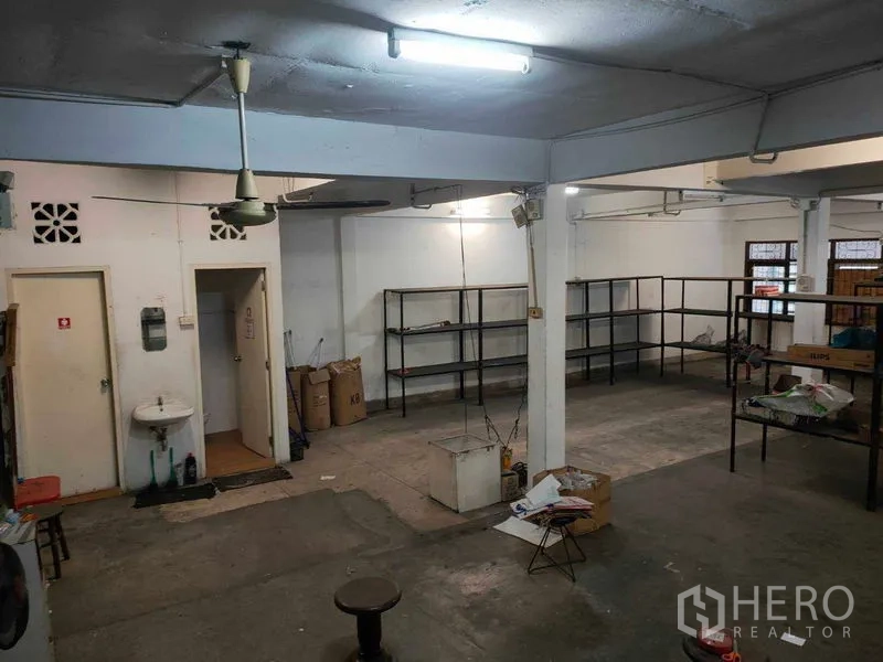Shophouse for rent in Watthana, Bangkok - Back-of-house storage area with concrete floor, ceiling fans, a sink and metal racks.