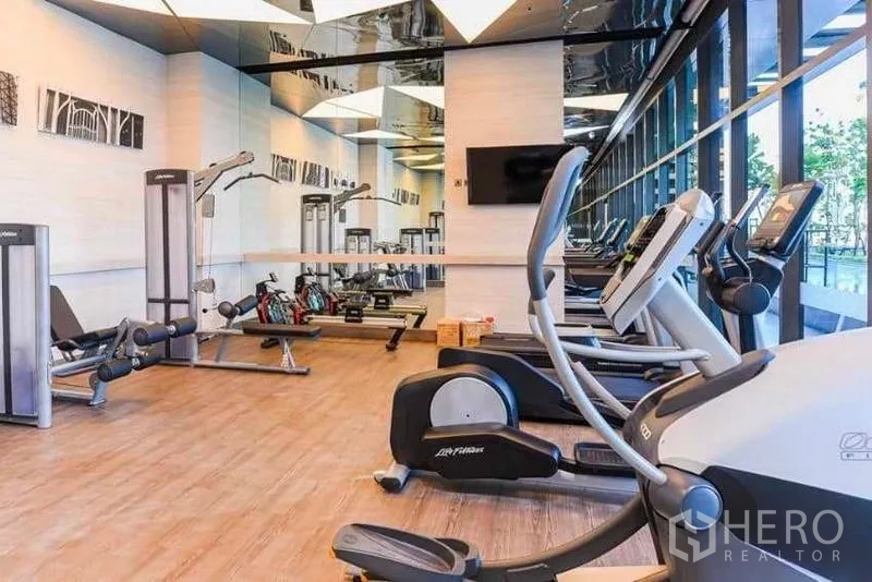 Condominium for rent in Phra Khanong, Bangkok - Condominium fitness center with treadmills, weight machines and a mirrored wall beside tall windows.