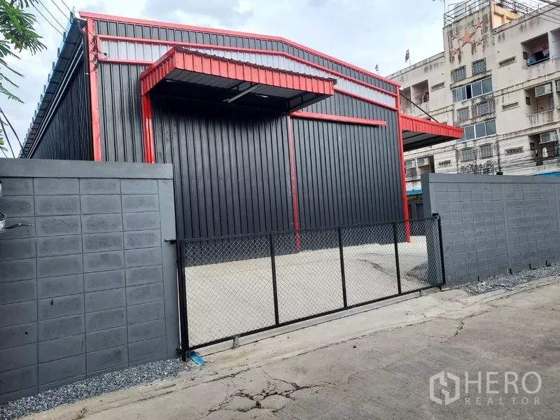 Warehouse for rent in Bang Kapi, Bangkok - Angled exterior of a steel warehouse with red trim, gable roof, chain-link gate and concrete forecourt beside low-rise buildings.