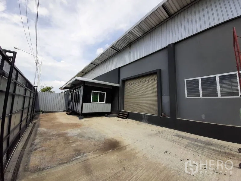 Factory for rent in Lam Luk Ka, Pathum Thani - Concrete yard with security gate, small site office and a large roller shutter loading bay.
