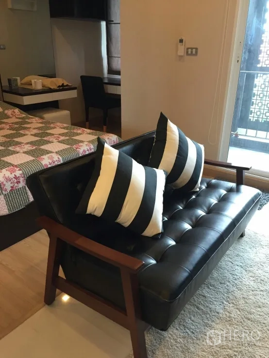 公寓 出租在Watthana, Bangkok - 客厅黑色皮质双人沙发配条纹靠枕，靠近阳台门与卧室区域。