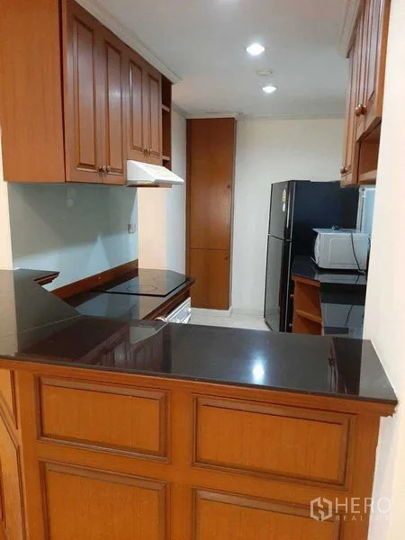 Apartment for rent in Watthana, Bangkok - U-shaped kitchen with wooden cabinetry, black stone countertops, cooktop with hood, fridge, and microwave.