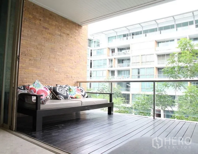 Condominium for rent in Khlong Toei, Bangkok - Covered balcony with sofa seating, glass railing and leafy outlook to neighboring residences.