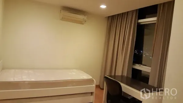 Condominium for rent in Khlong Toei, Bangkok - Second bedroom with single bed, study desk by a large window with city night view and air conditioner.