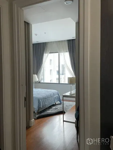 Condominium for rent in Khlong Toei, Bangkok - Cozy bedroom with a large window, sheer curtains, soft blue bedding, and wood flooring.