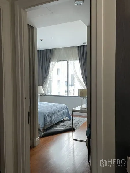 Condominium for rent in Khlong Toei, Bangkok - Cozy bedroom with a large window, sheer curtains, soft blue bedding, and wood flooring.